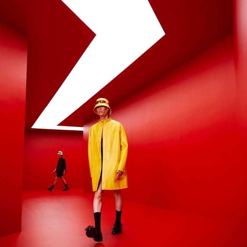 Seorang model bermantel kuning berjalan melalui terowongan merah