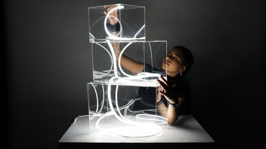 A person holds three stacked boxes on top of a podium containing illuminated ribbons of light