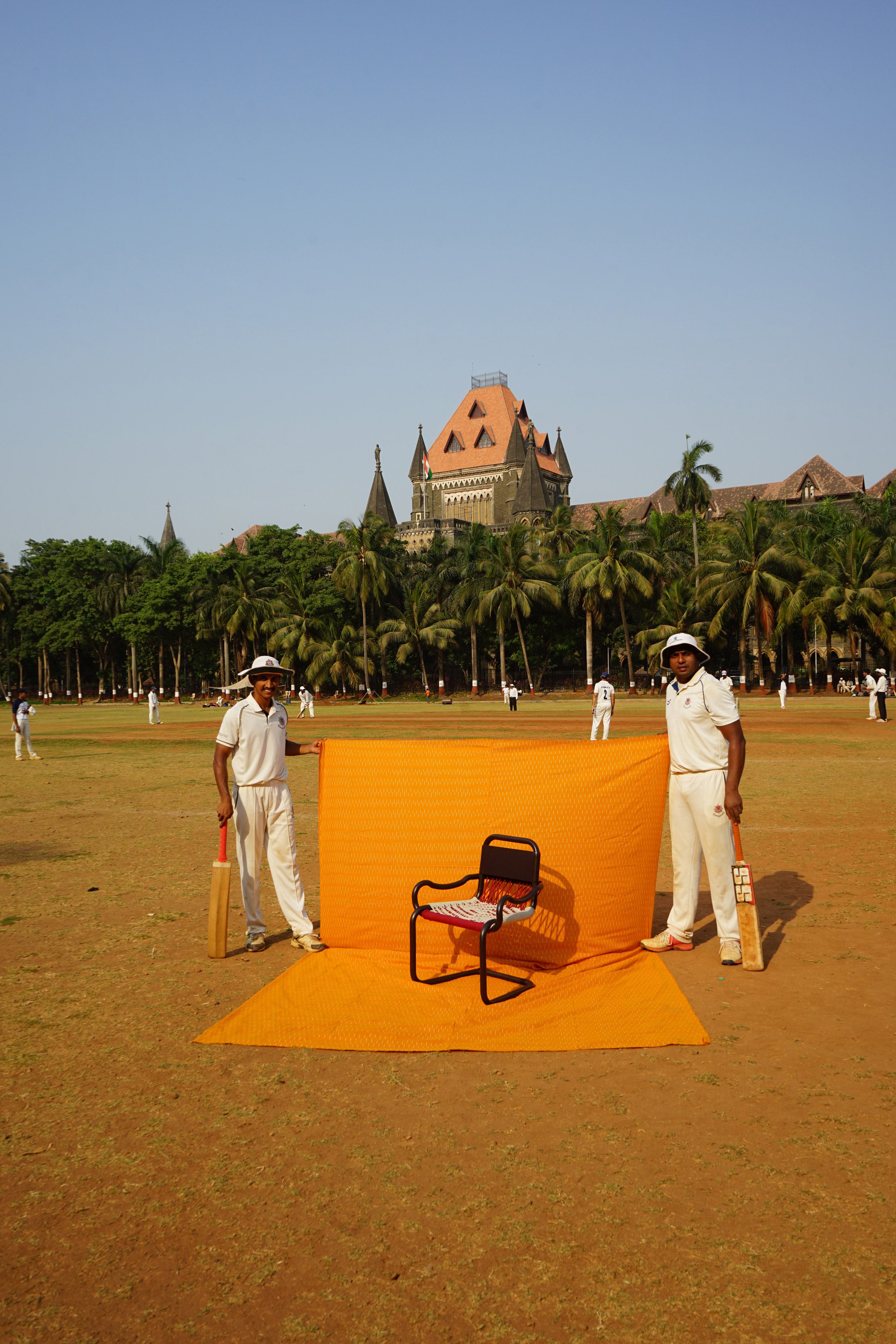 Chair photographed against a yellow sheet held by two cricketers on a cricket field in India