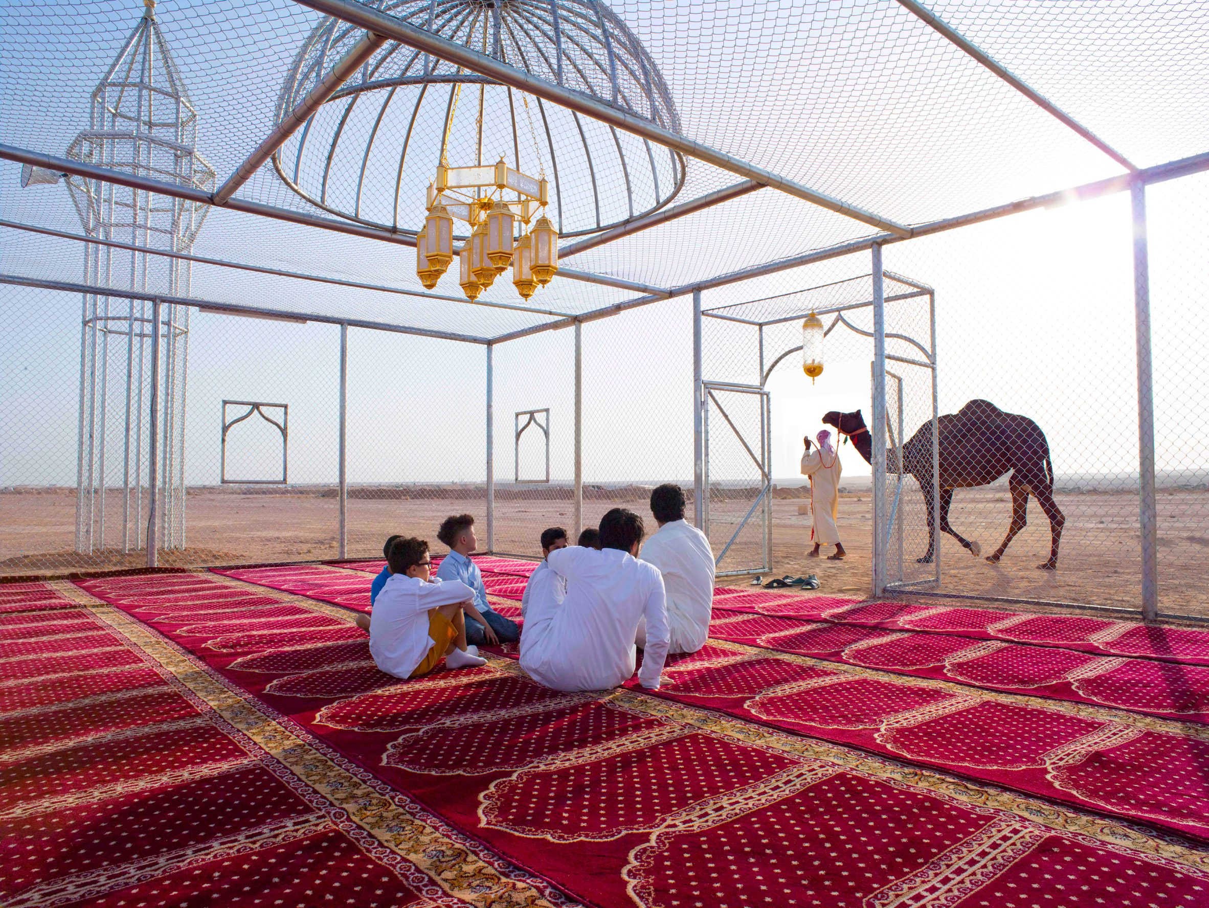 A group of people sit under the dome of the wire mosque, looking out at a passing camel