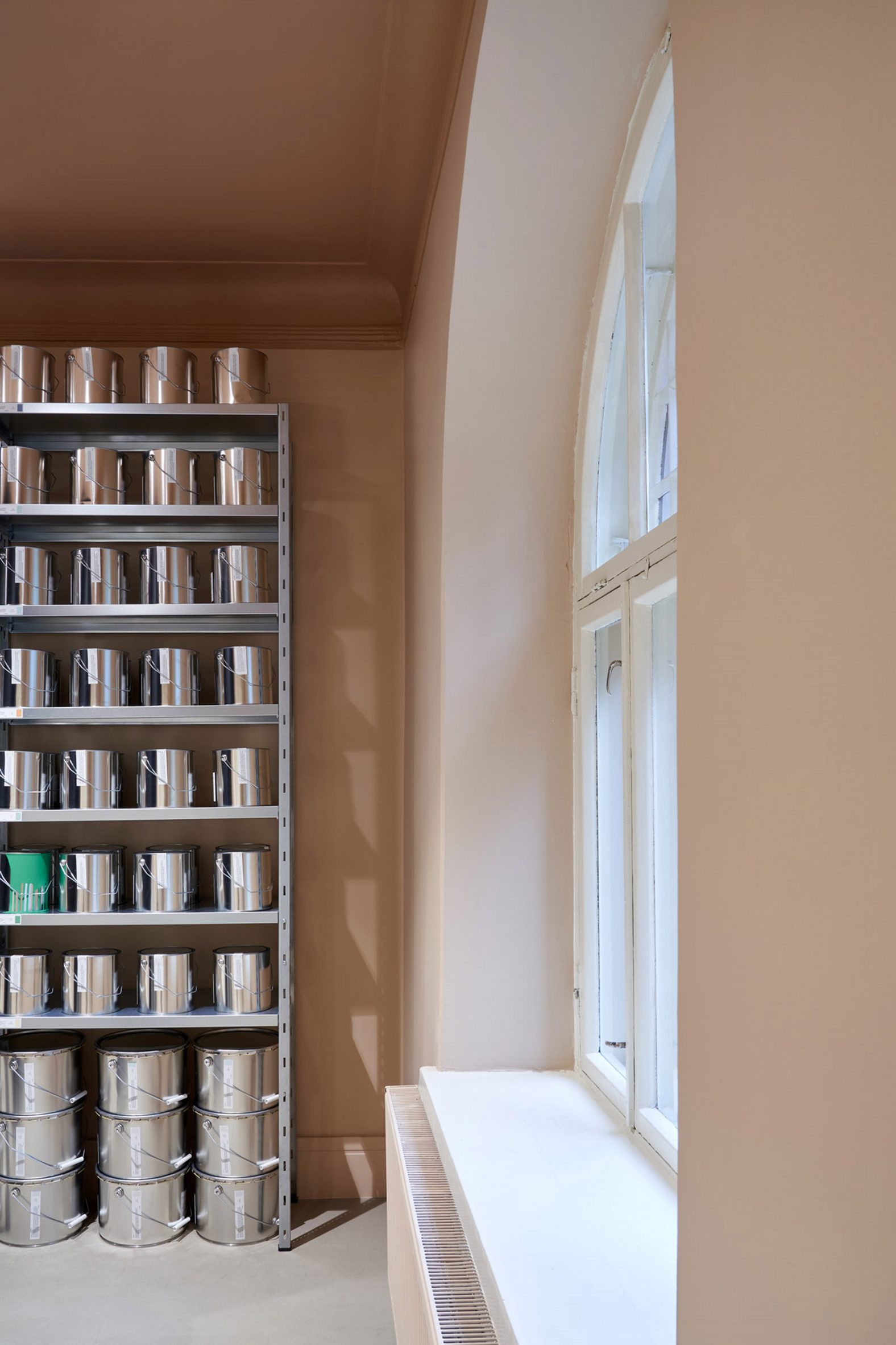 Shelves of paint tins in a pale pink room lit by a large arched window in Cover Story's shop