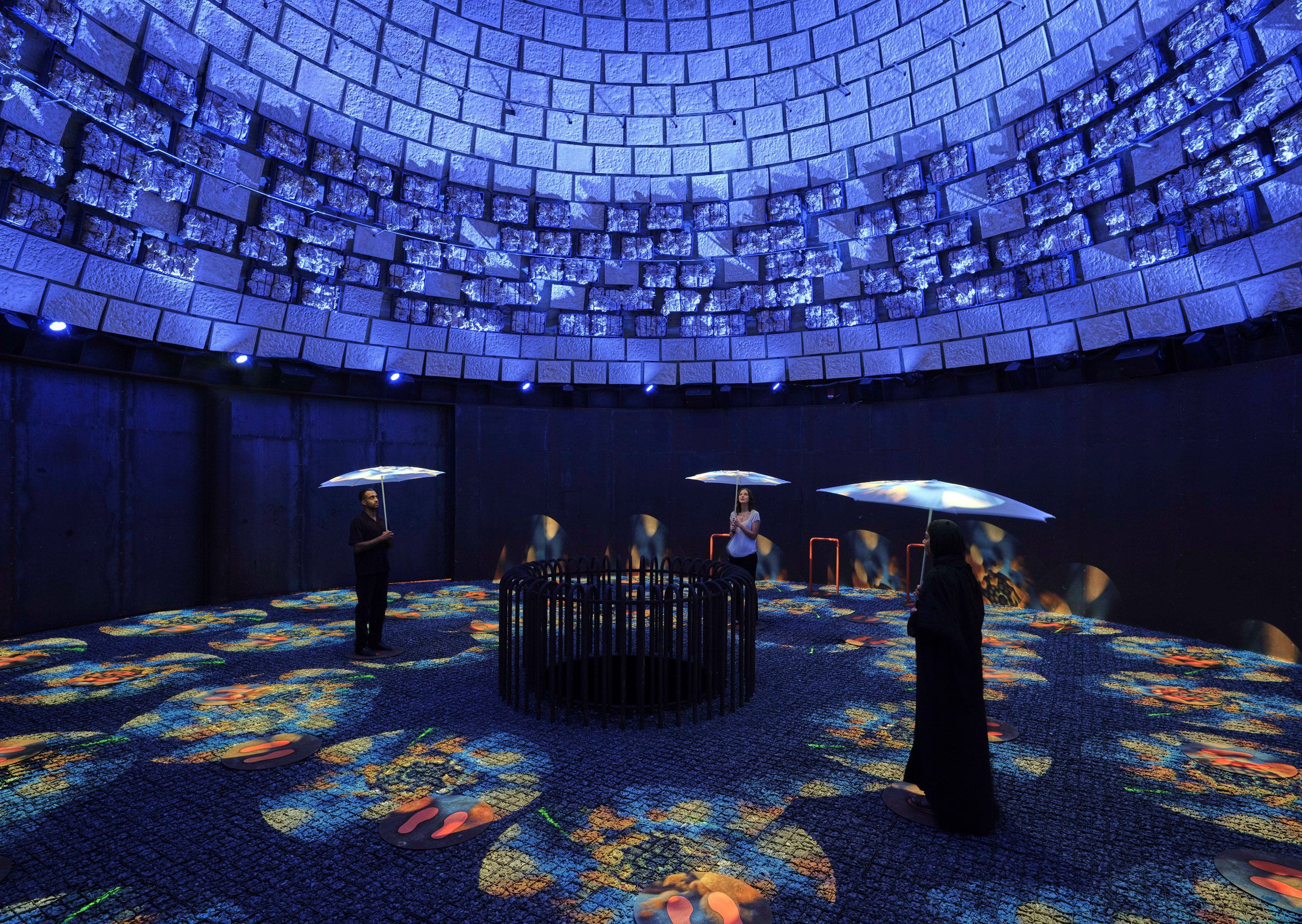 People with umbrellas stand in a dark, circular space with projections on the floor