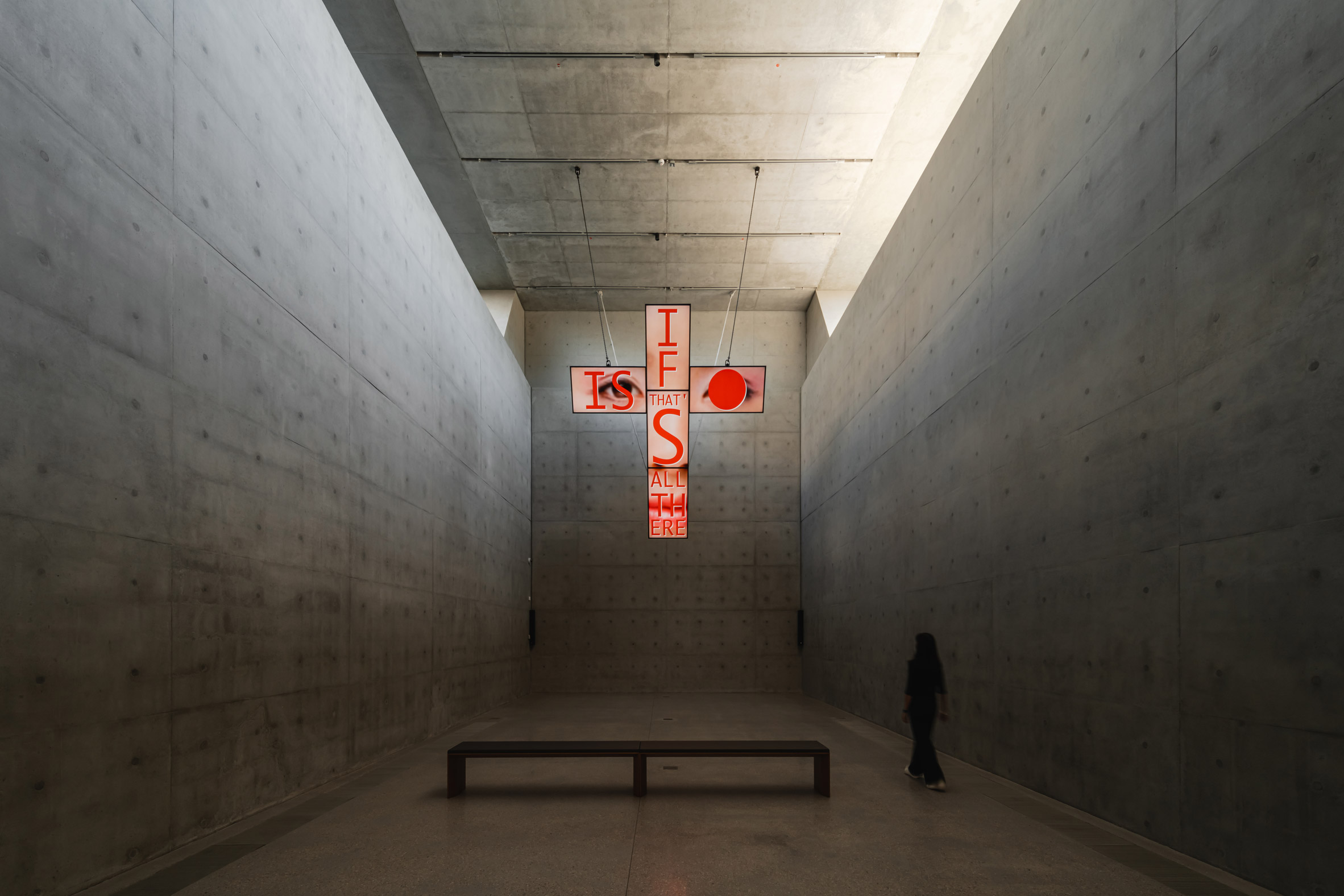 Illuminated cross-shaped artwork hangs high up in an all-concrete gallery space