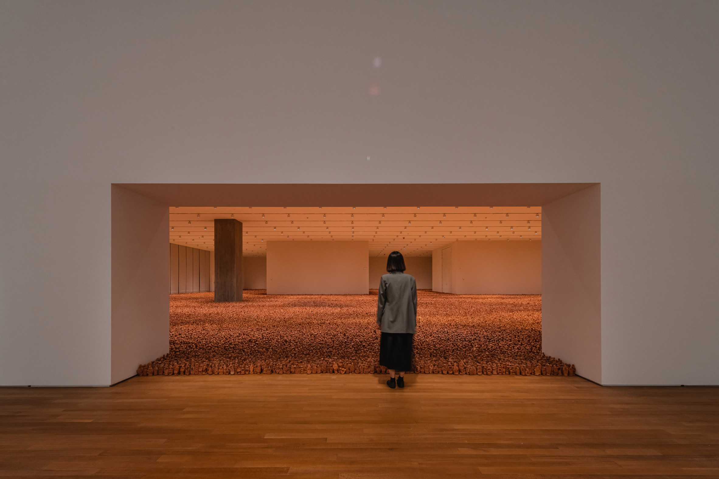Thousands of small clay figurines stare outwards from a large white gallery space towards one watchful visitor