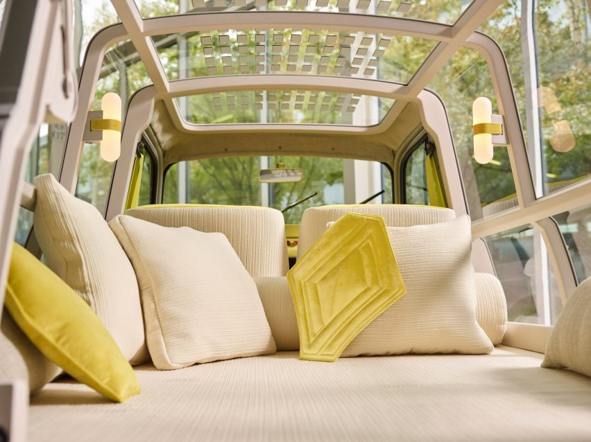 Bed in the boot of a car with cream chenille upholstery, cushions and wraparound transparent panelling showing views of a forest