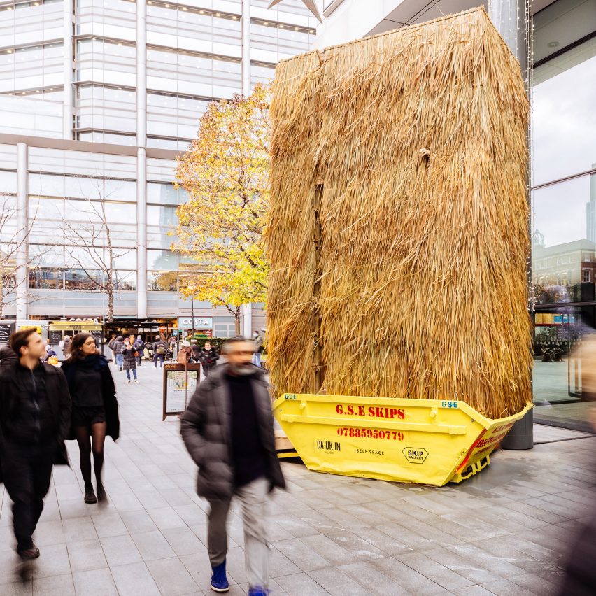 People walk passed a grass thatched installation in a skip