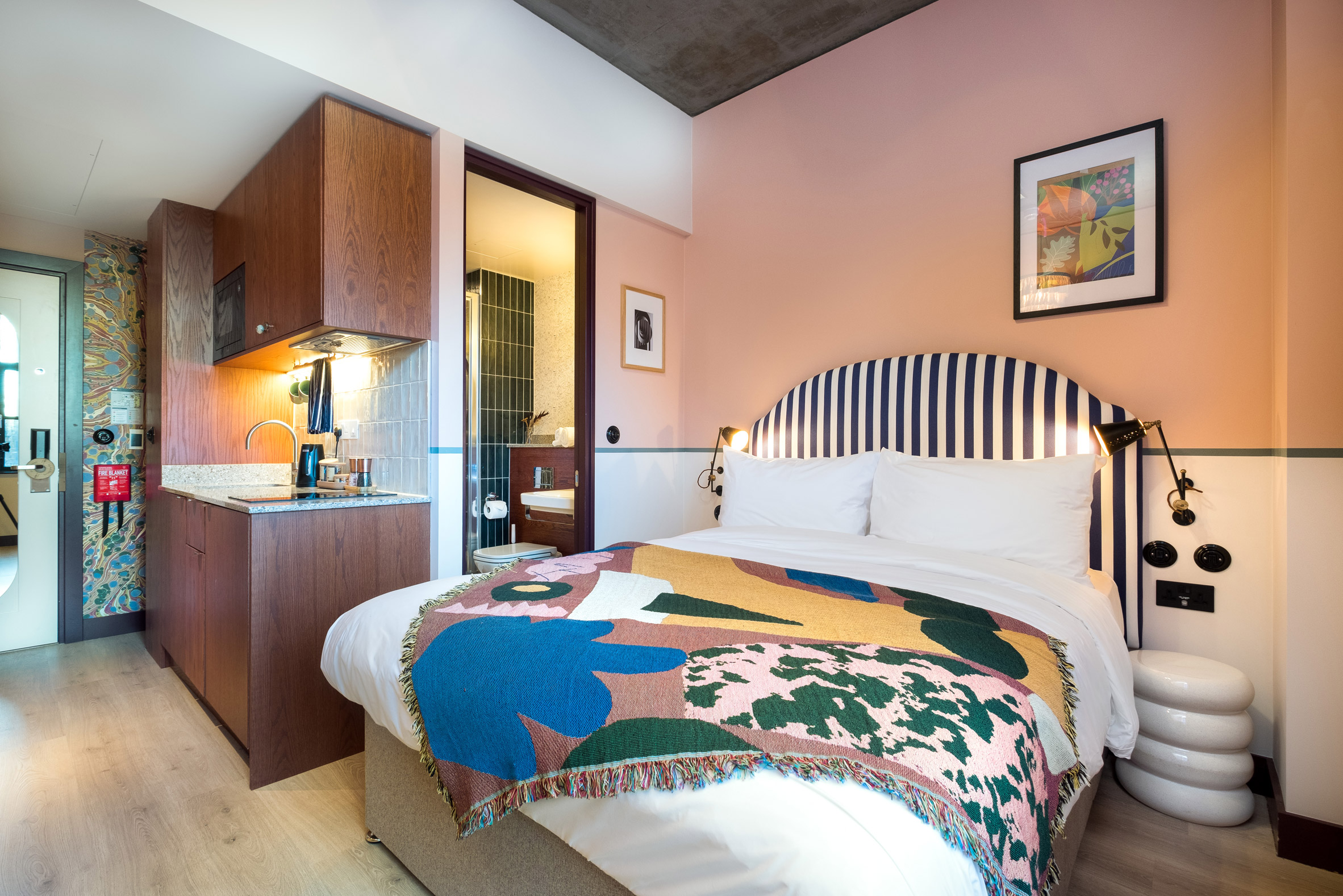 Hotel room with wooden kitchenette, pink walls and colourful throw over double bed
