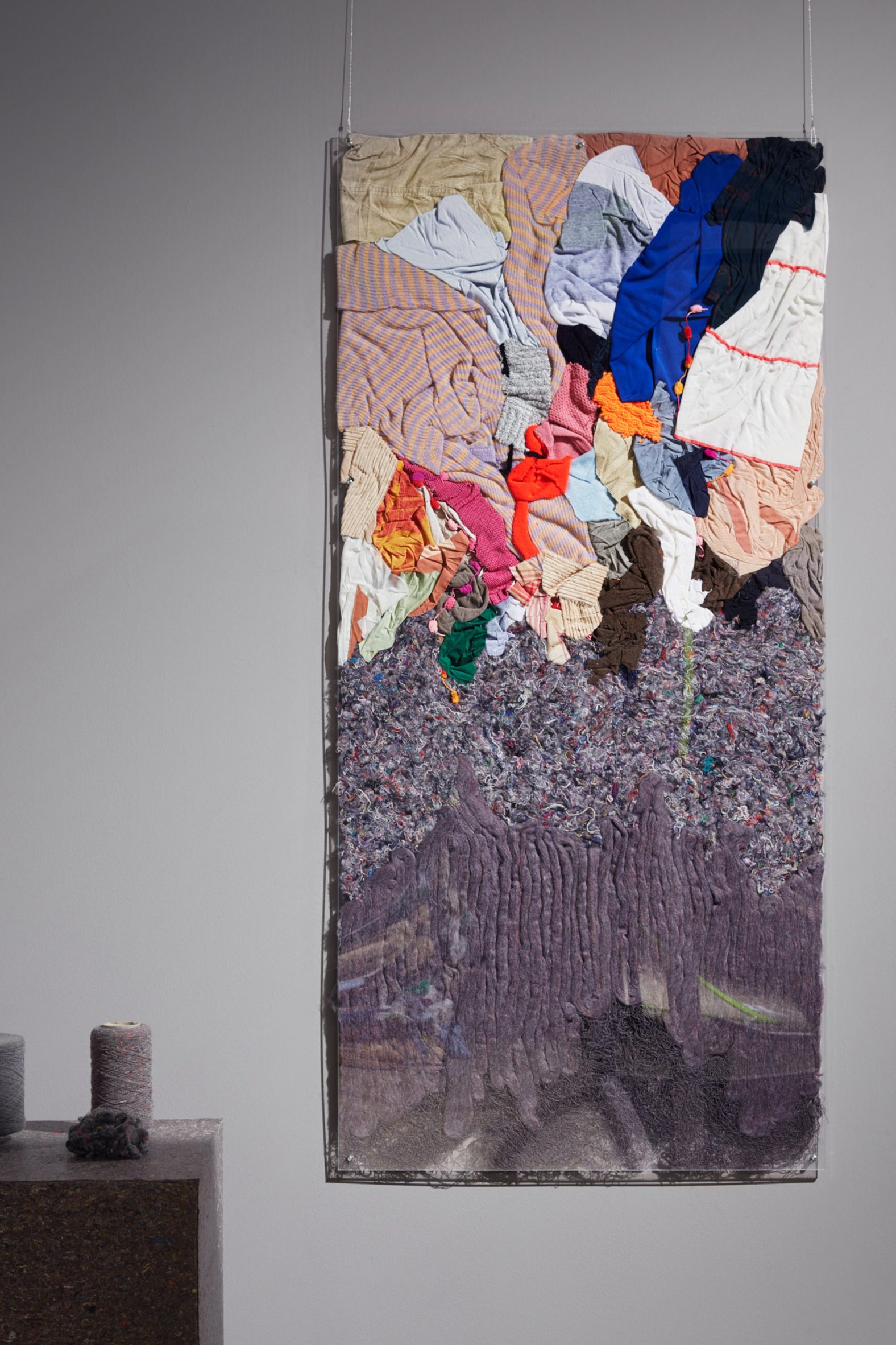 Wall hanging showing mixed garments at the top, unravelling into a grey mix of fibres and then becoming yarn