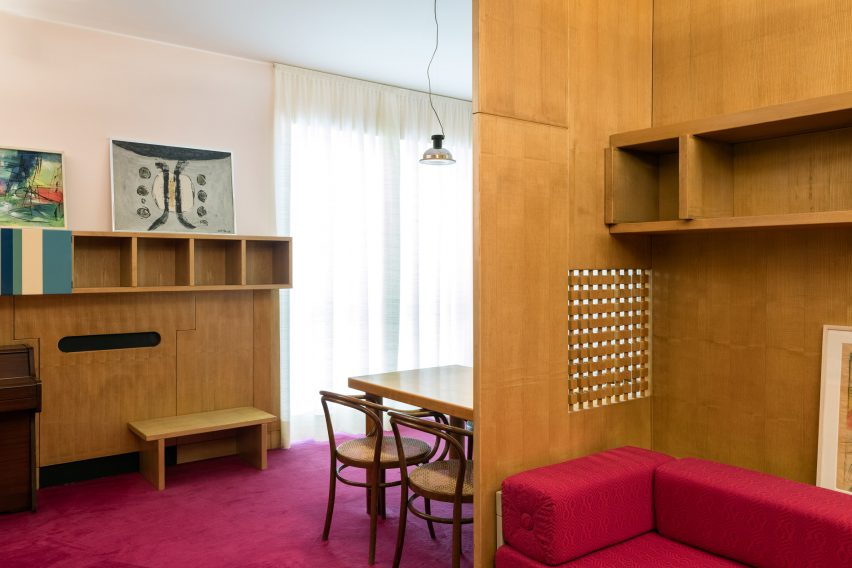 Dining area with wooden chairs surrounded by built-in wooden shelving and magenta carpet in living room of the Casa Lana installation at Triennale di Milano, Italy