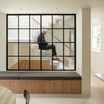 Man sitting on a staircase behind a wall of gridded glazing inside Danish Mews House by Neil Dusheiko Architects