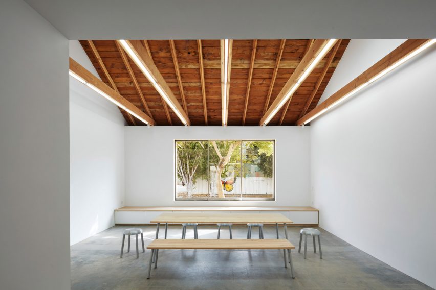 Open white-walled exhibition and event space with a long row of benches and seating and a large window looking onto the garden at Emeco House