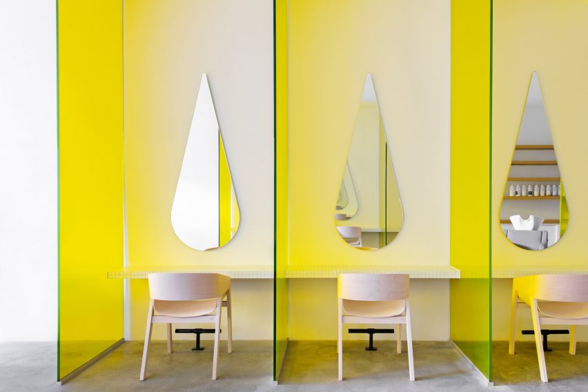 Yellow tinted glass partitions separate hair cutting stations at a hair salon, with pale timber chairs and teardrop-shaped mirrors in each station