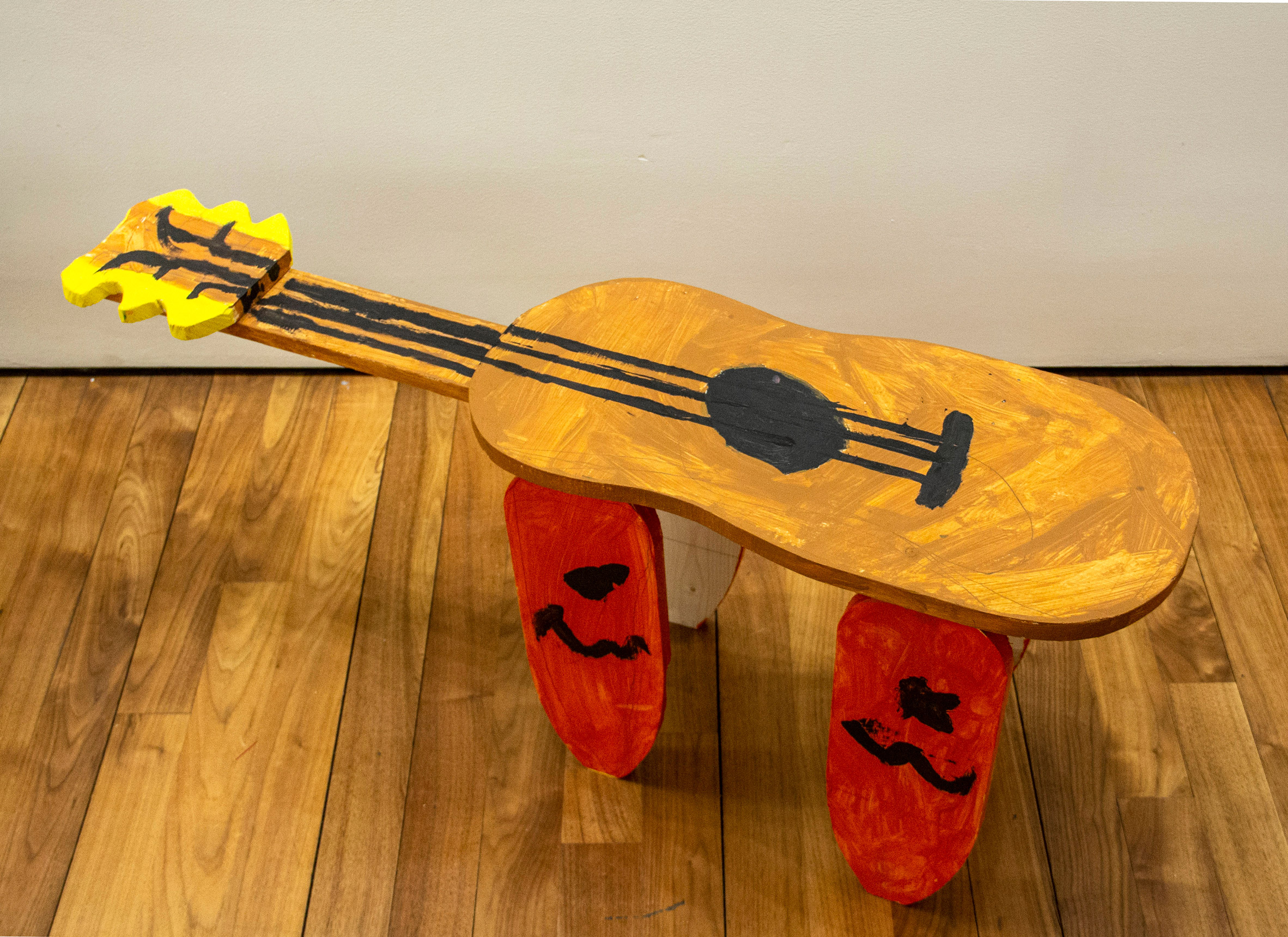 Child-made bench with seat in the shape of a guitar and four legs in the shape of red guitar picks 