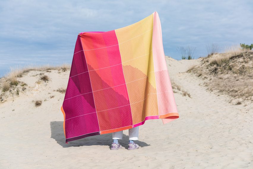 Person on sand dune holding up bright red, purple, orange and yellow blanket with patterns of lines