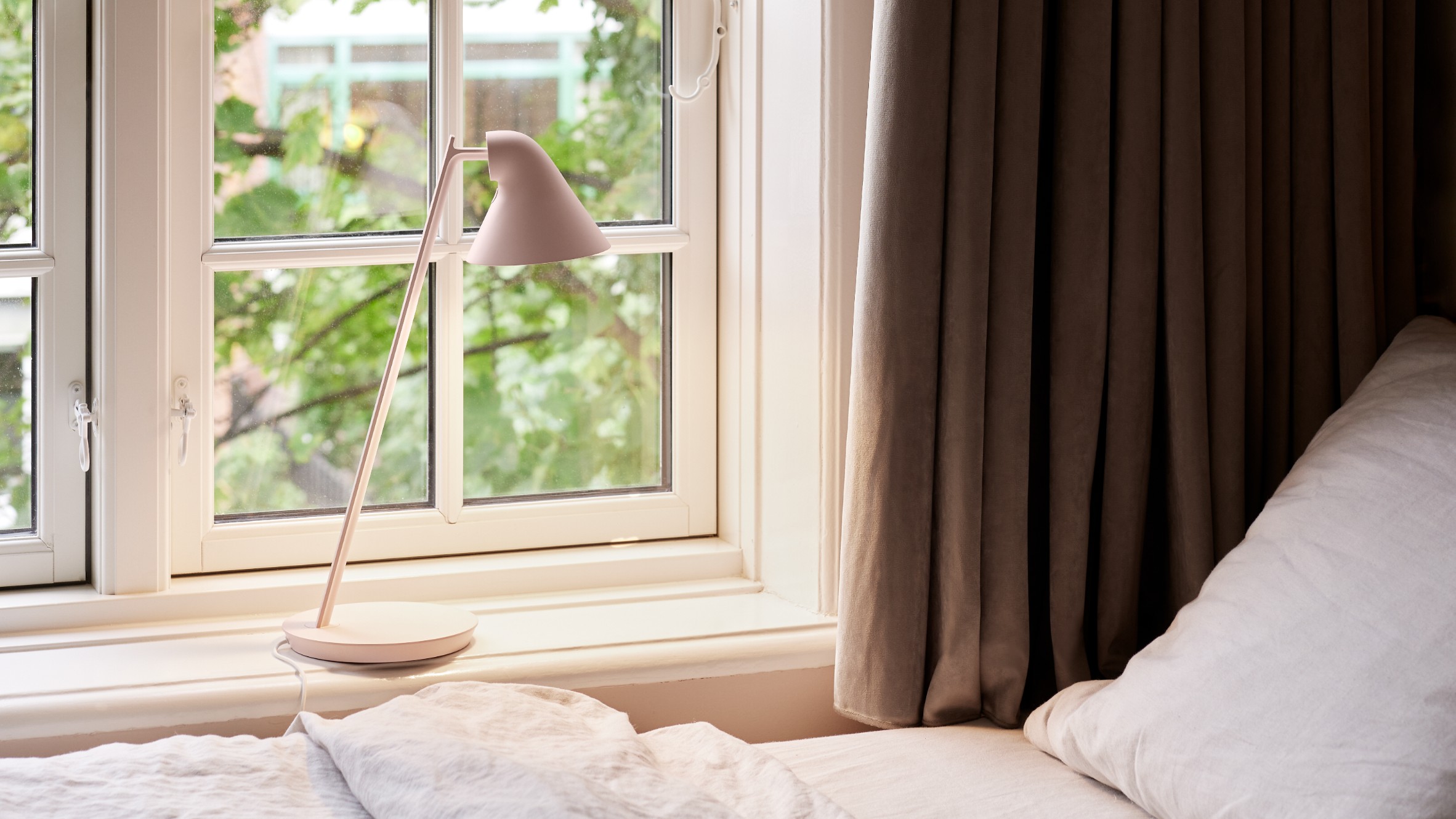 A photograph of the NJP Mini table lamp by Louis Poulsen in soft pink stands on a bedroom windowsill