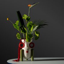 White Explorer vase with green leg stands and red handles in a showroom space with large green leafs and flowers