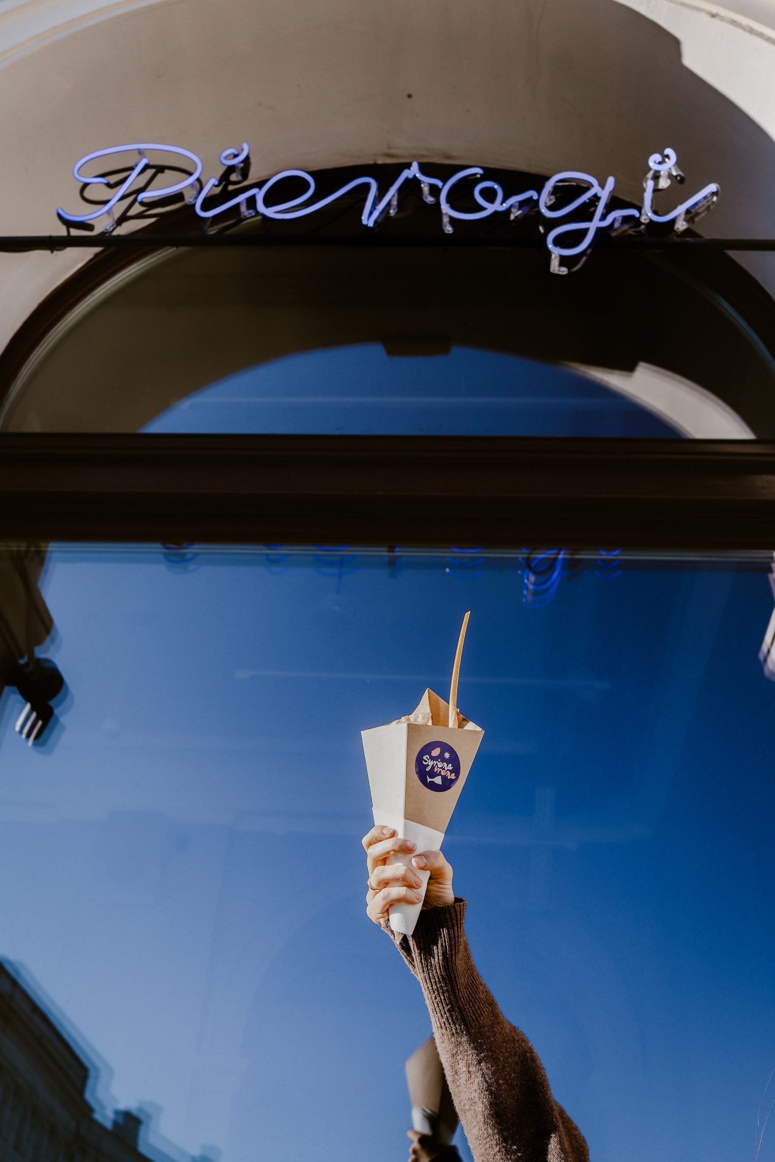Exterior Syrena Irena pierogi restaurant with neon sign and hand holding a cone of dumplings