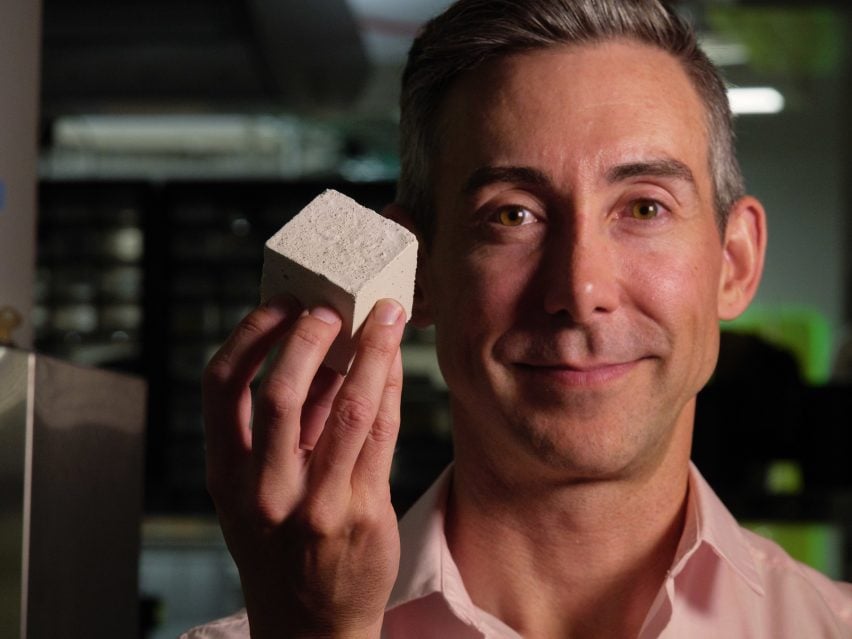 Wil Srubar faces camera holding a cube of concrete made with algae-grown limestone
