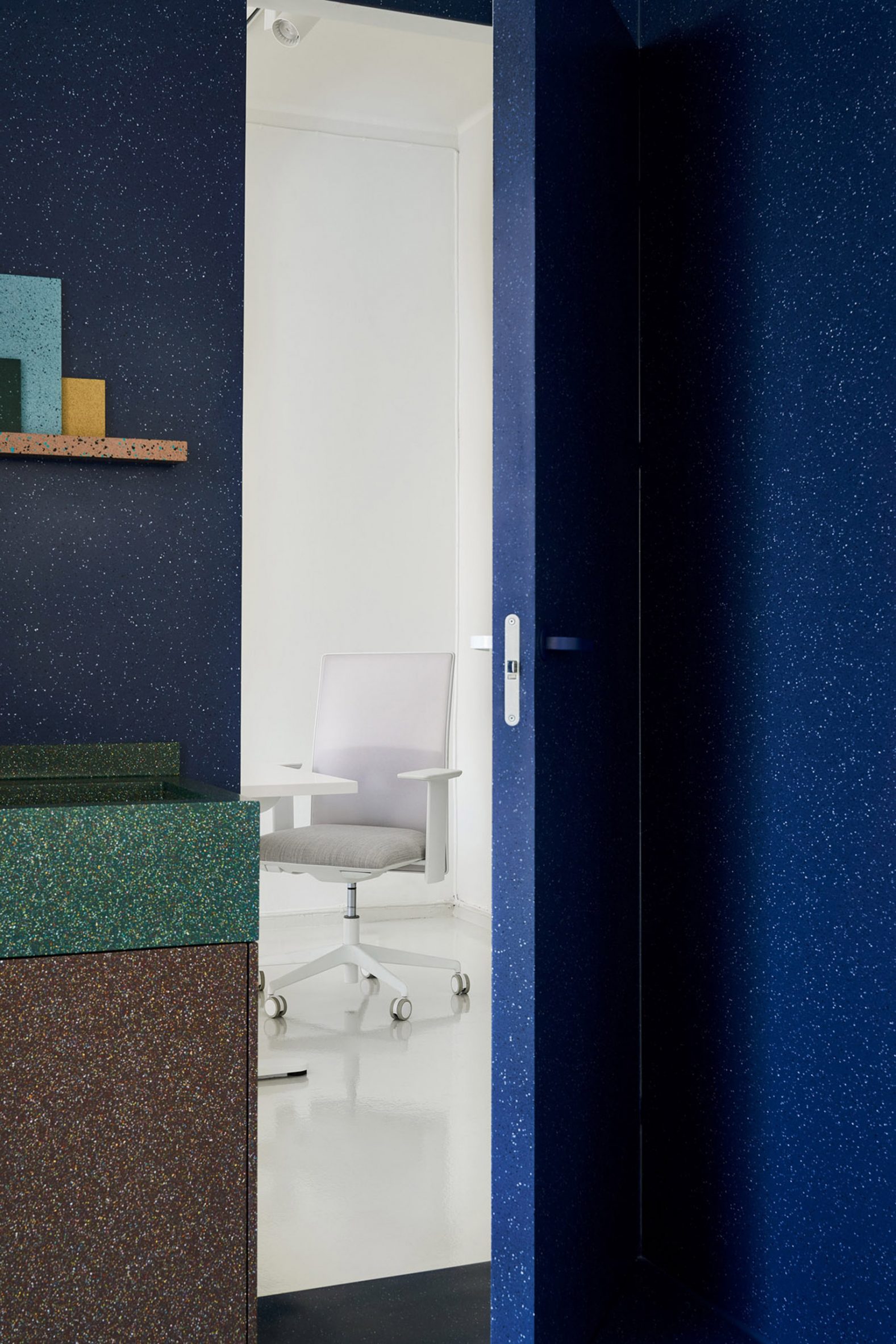Door open in a dark blue wall created with Durat surface material reveals a white office area behind it