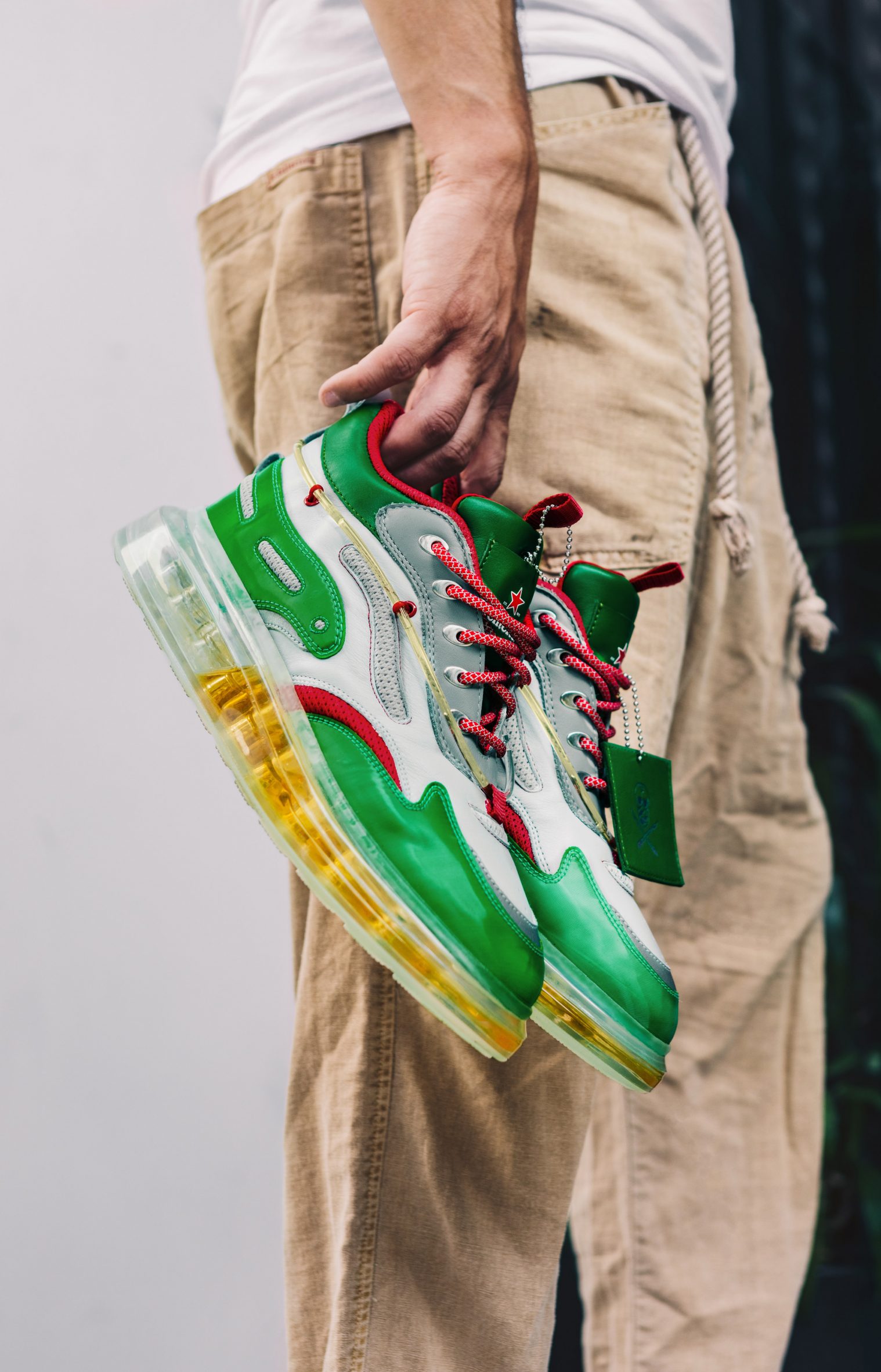 A photograph of someone holding a pair of red, white and green Heinekicks sneakers with a transparent sole filled with beer