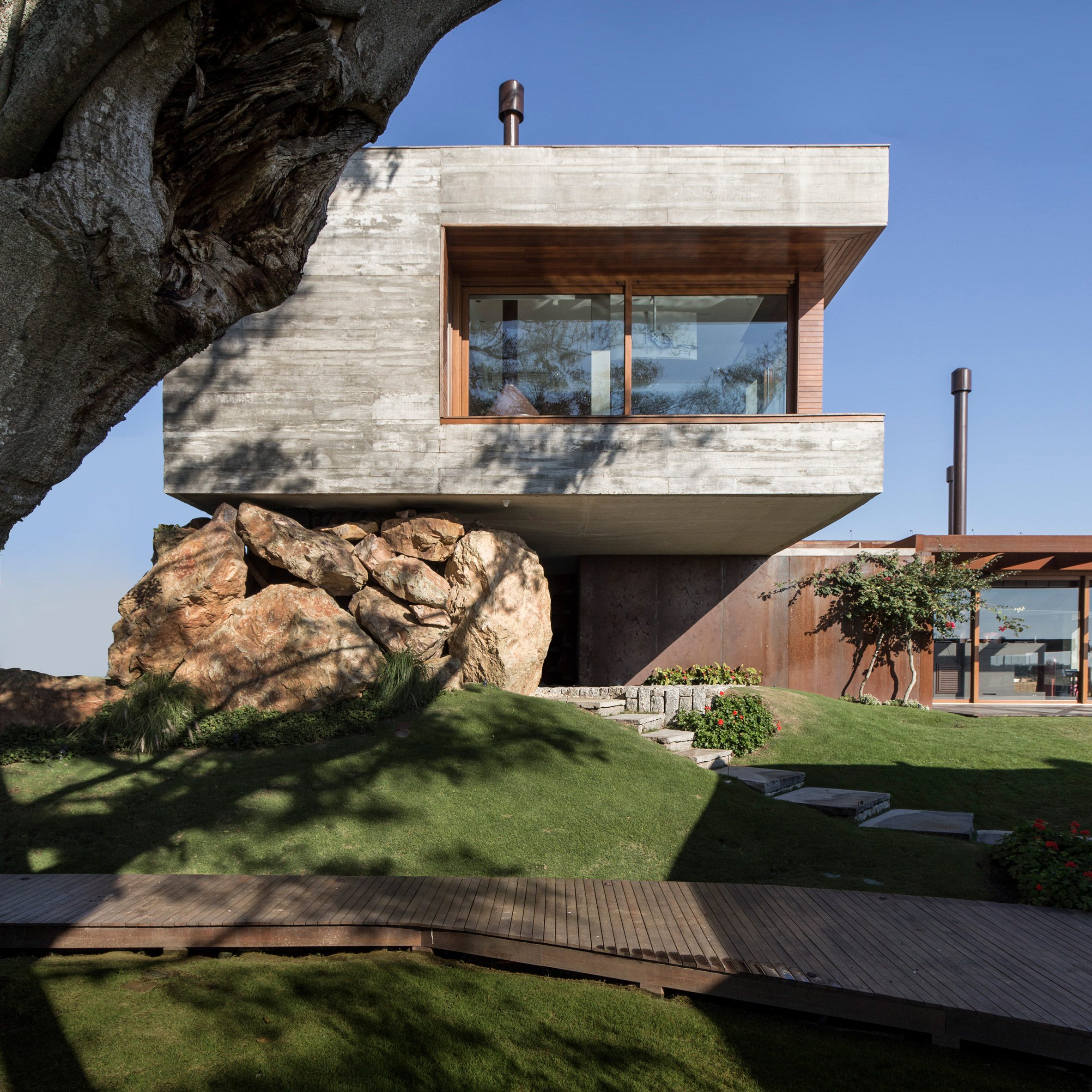 Concrete house with window cut-out cantilevering over a grass garden and balanced on a large rock