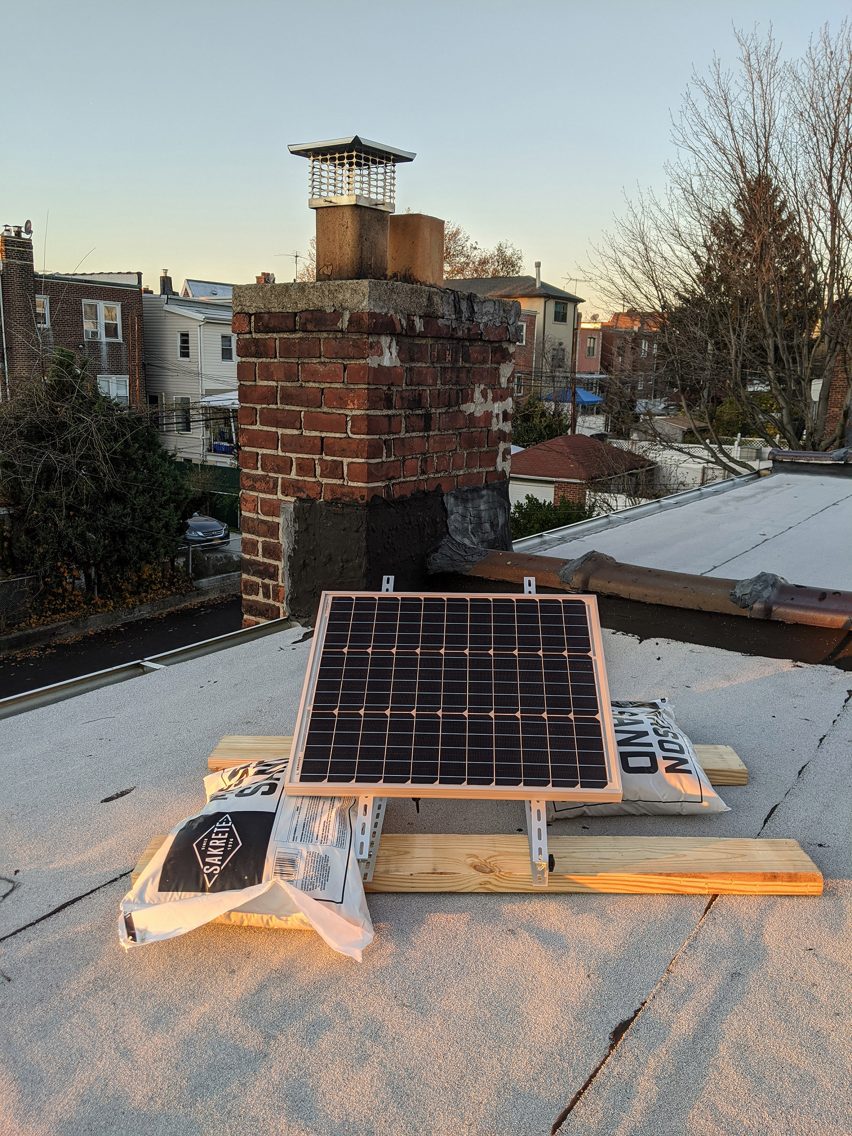 A solar panel sits on a rooftop, held in place by sand bags