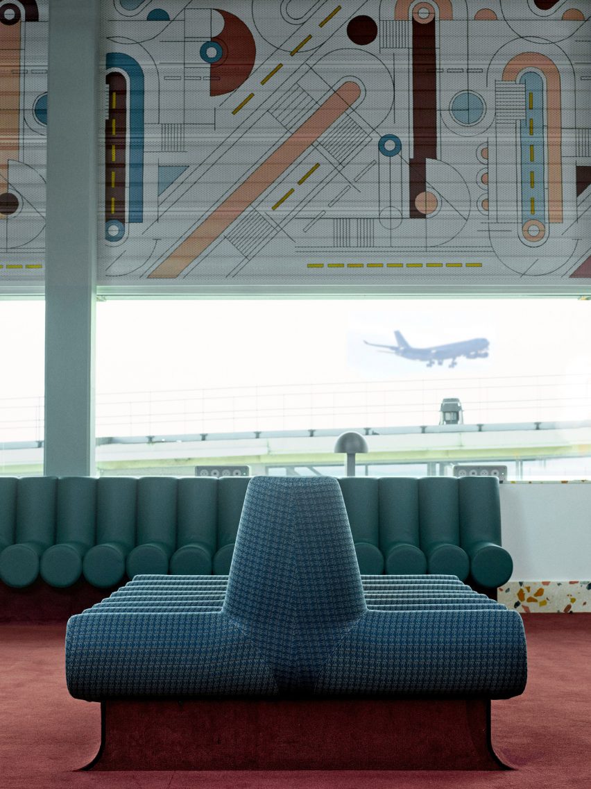 Фотография, показывающая зал вылета аэропорта с абстрактной фреской над окнами и зелеными сиденьями.