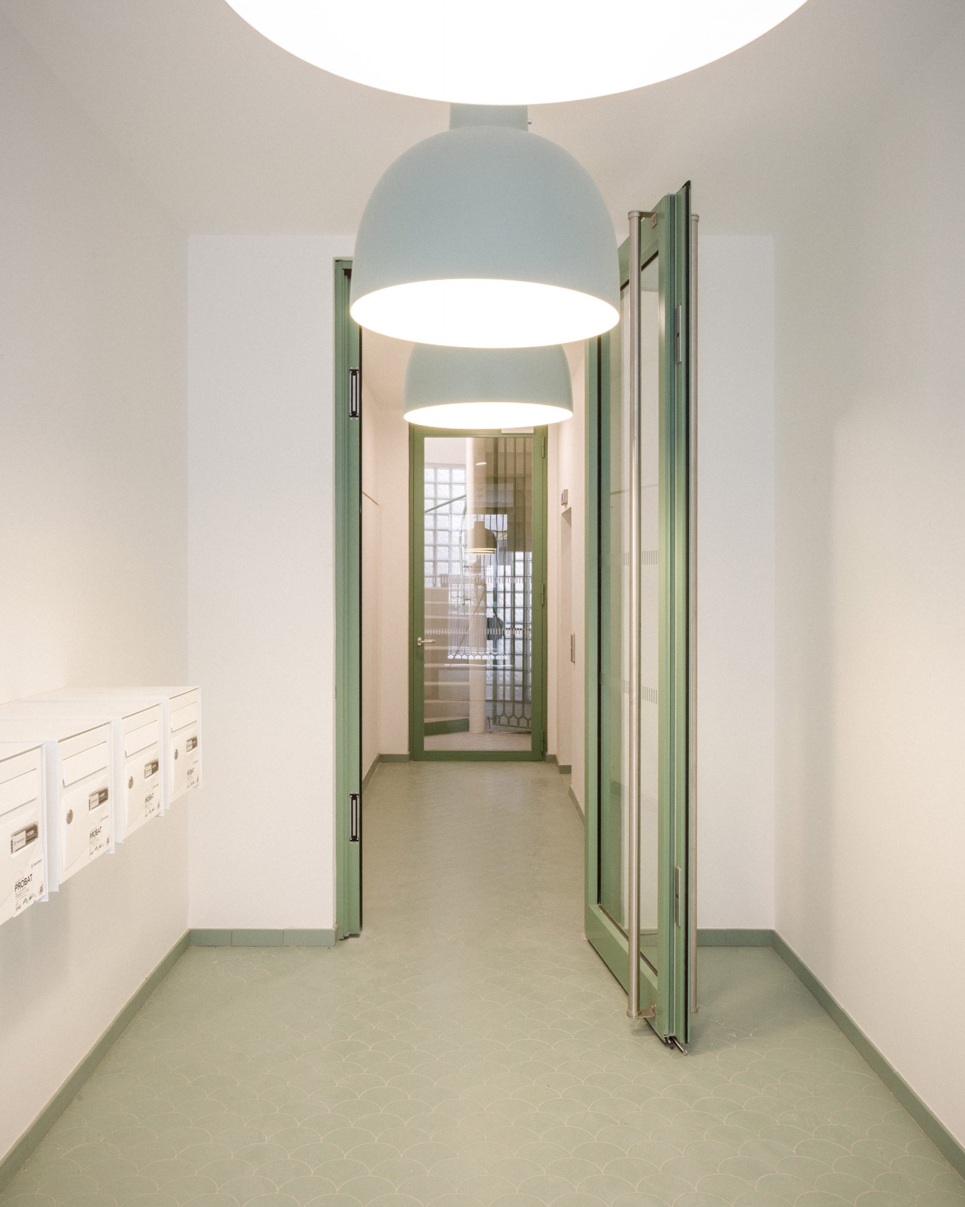 Lobby of Jean Christophe Quinton's Paris social housing project with white postboxes on the left, soft green ogee tiles on the floor, green door frames and skirting boards and paler green overhead lamp shades