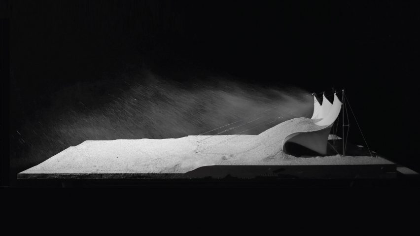 Photograph of a tabletop model with sand blowing against a structure on the right hand side, in front of a dark backdrop
