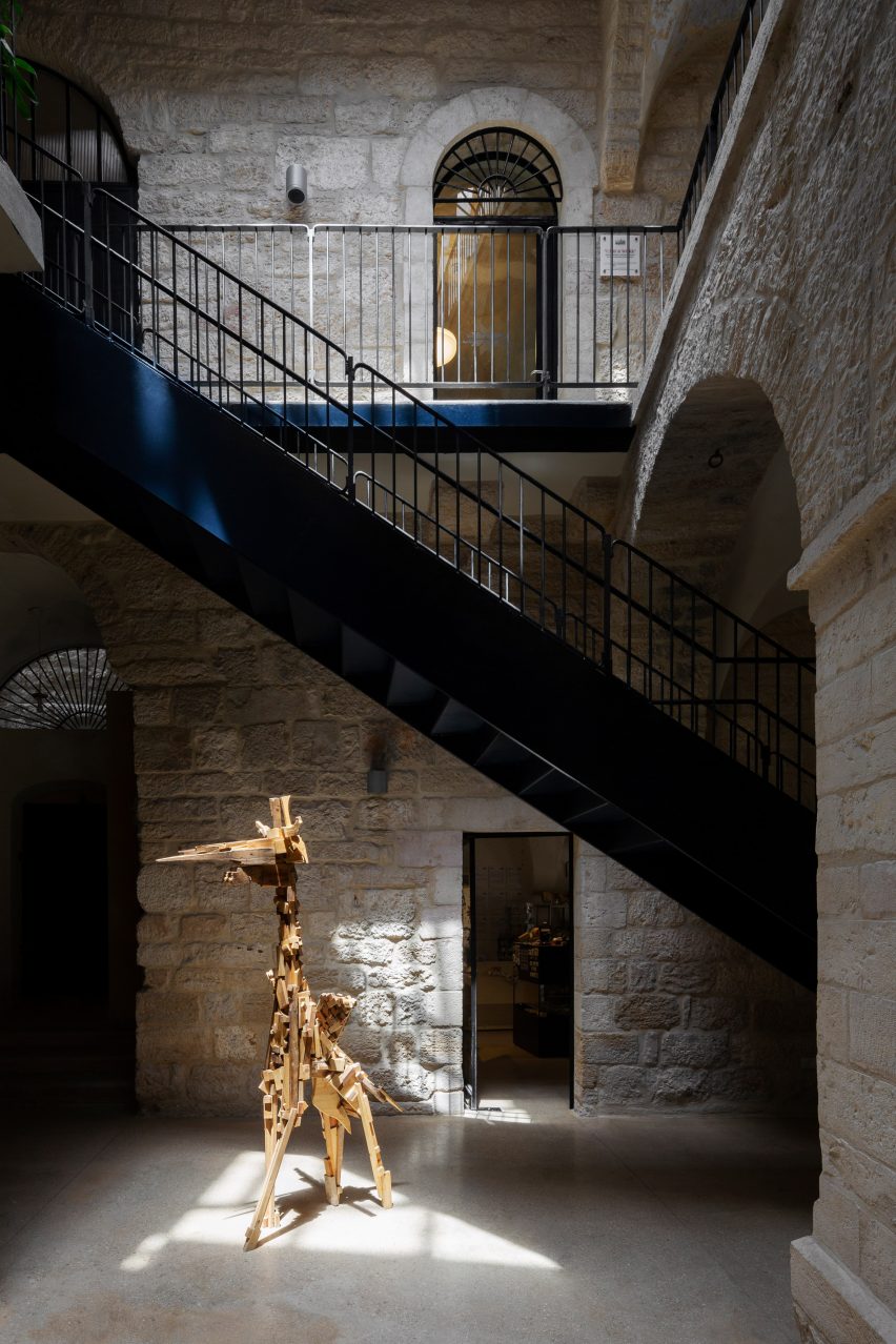Black steel staircase cutting through a stone courtyard with a giraffe-like sculpture in it