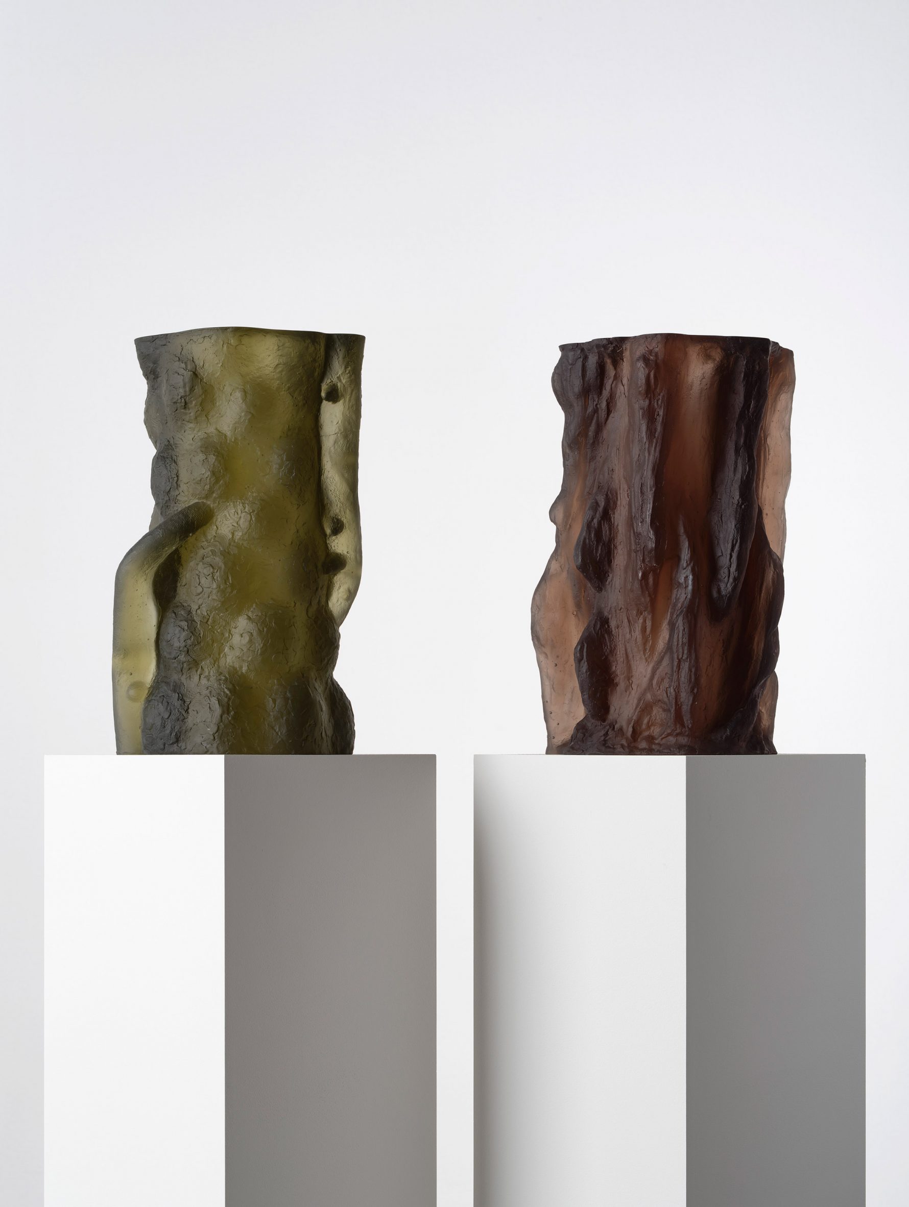 Photograph showing a green and brown-toned textural glass sculptures on white plinths
