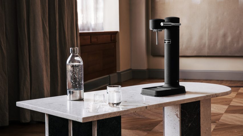 Photo of the Aarke Carbonator 3 in Matte Black finish sitting on a white table with a bottle and glass of sparkling water