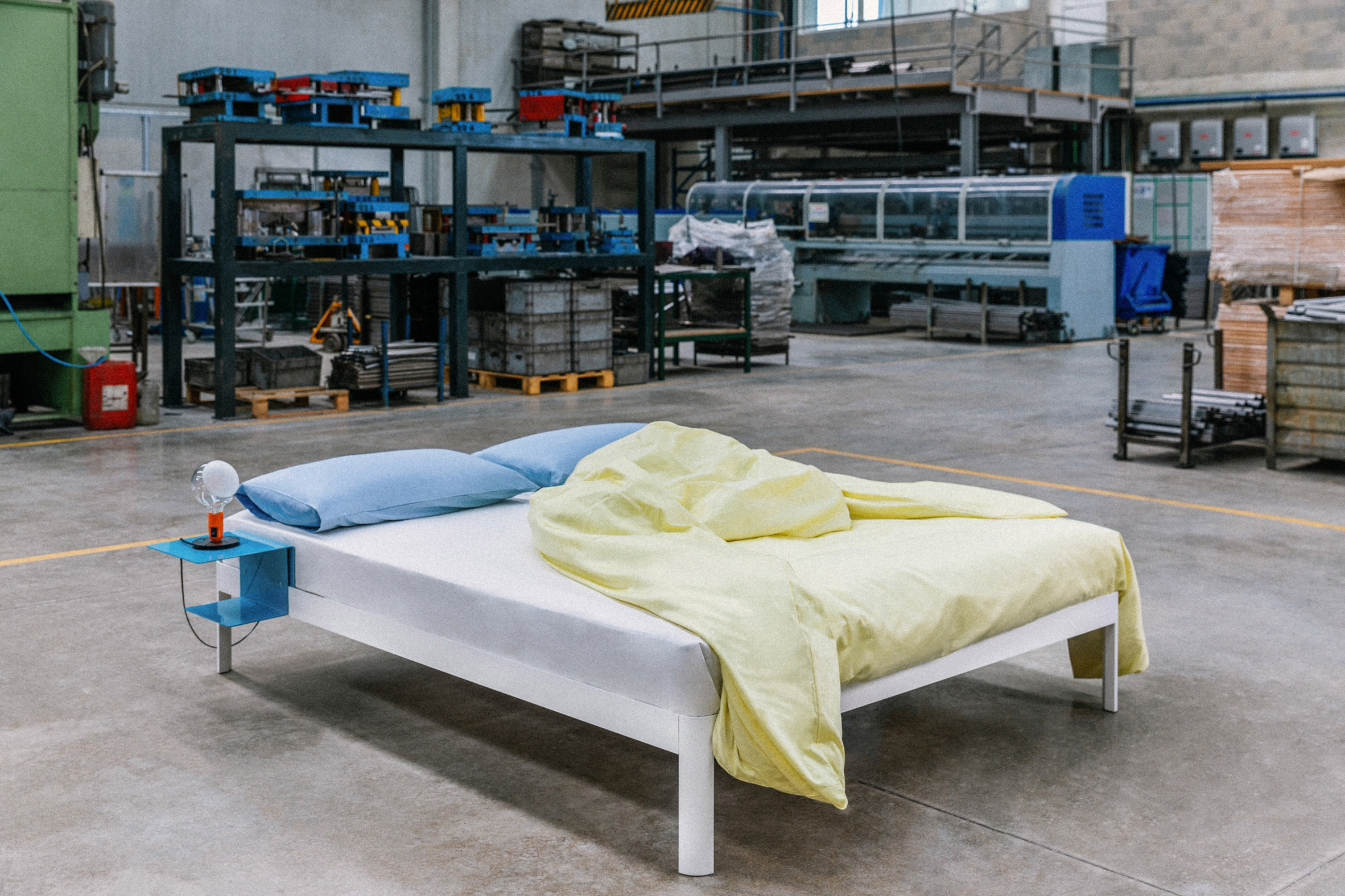 Photograph of a white bed with yellow and blue bed sheets in an industrial factory