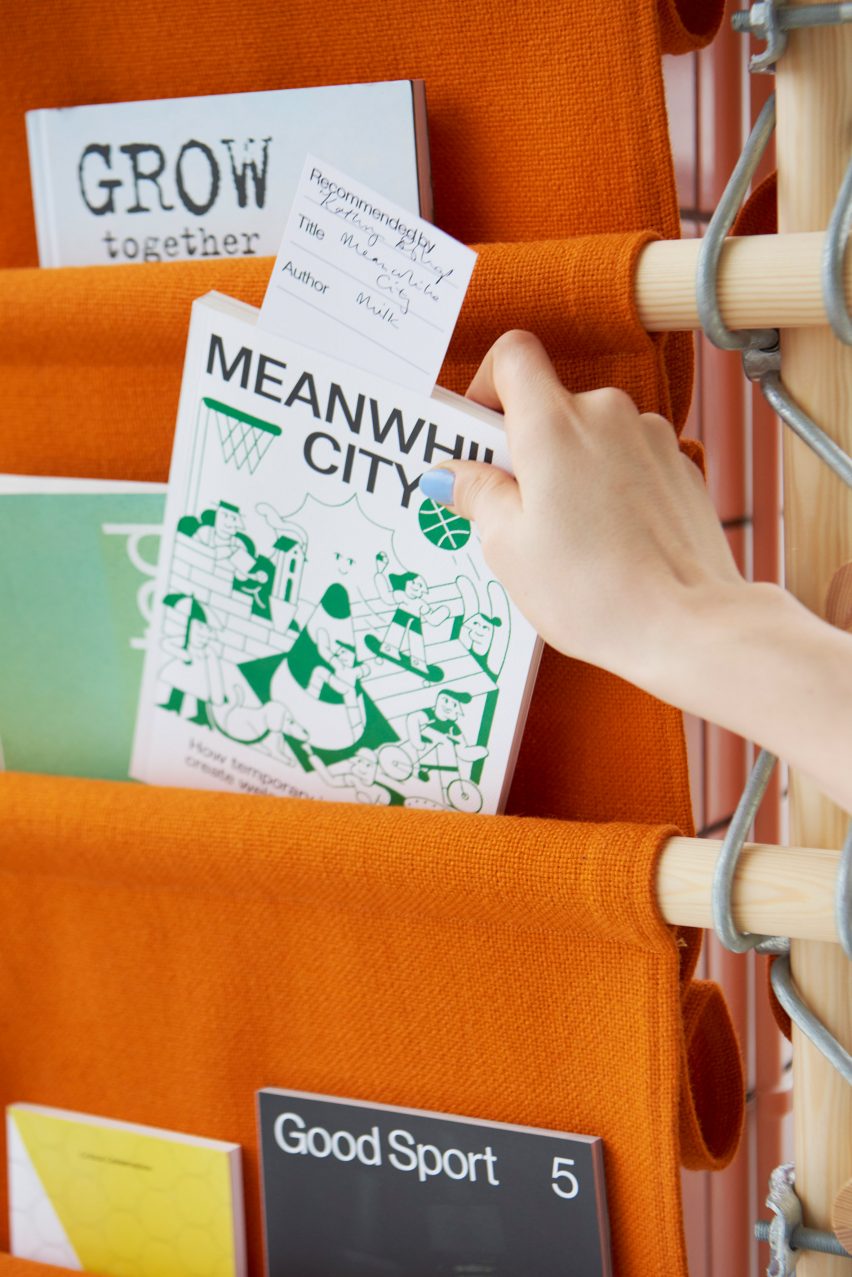 Detailní fotografie ruky beroucí knihu s názvem Mezitím město z jasně oranžového látkového stojanu