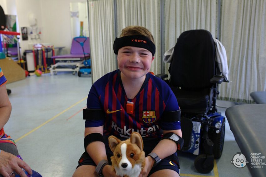 Photo of a child wearing sensors strapped to their limbs with Velcro and smiling at the camera while holding a stuffed Corgi toy
