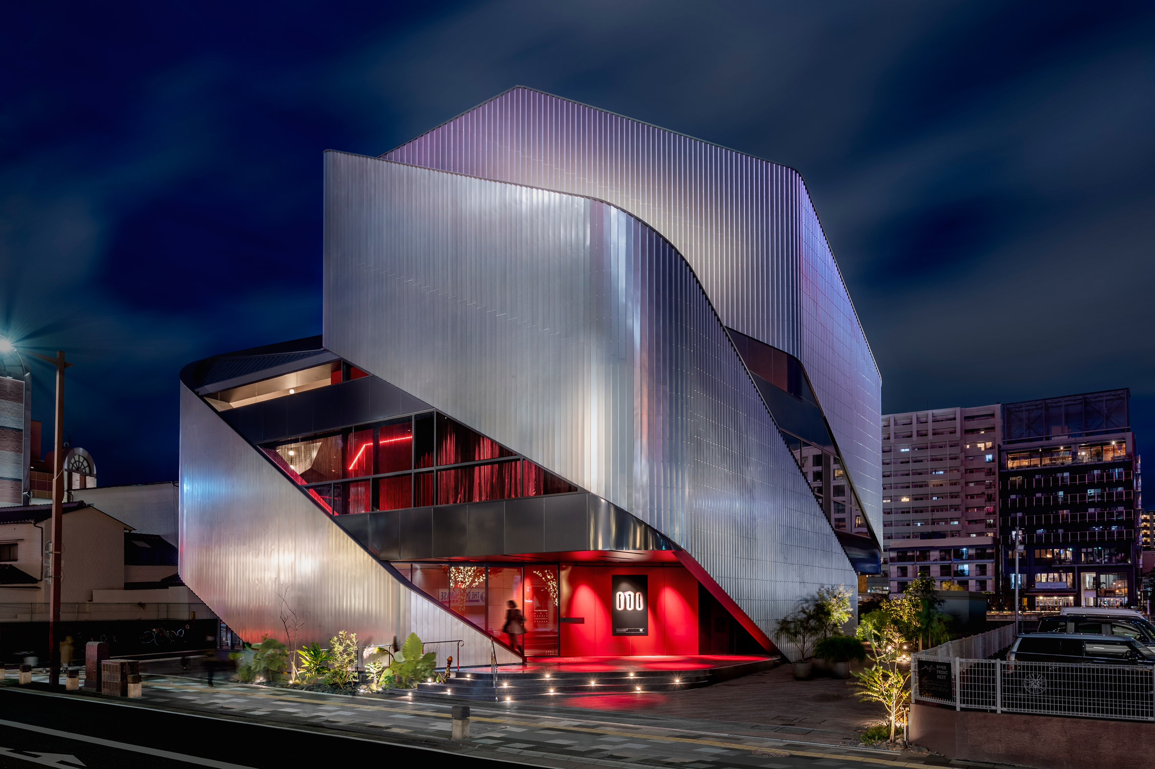 A theatre building on a city street corner with sweeping steel exterior panels lit by spotlights on the ground