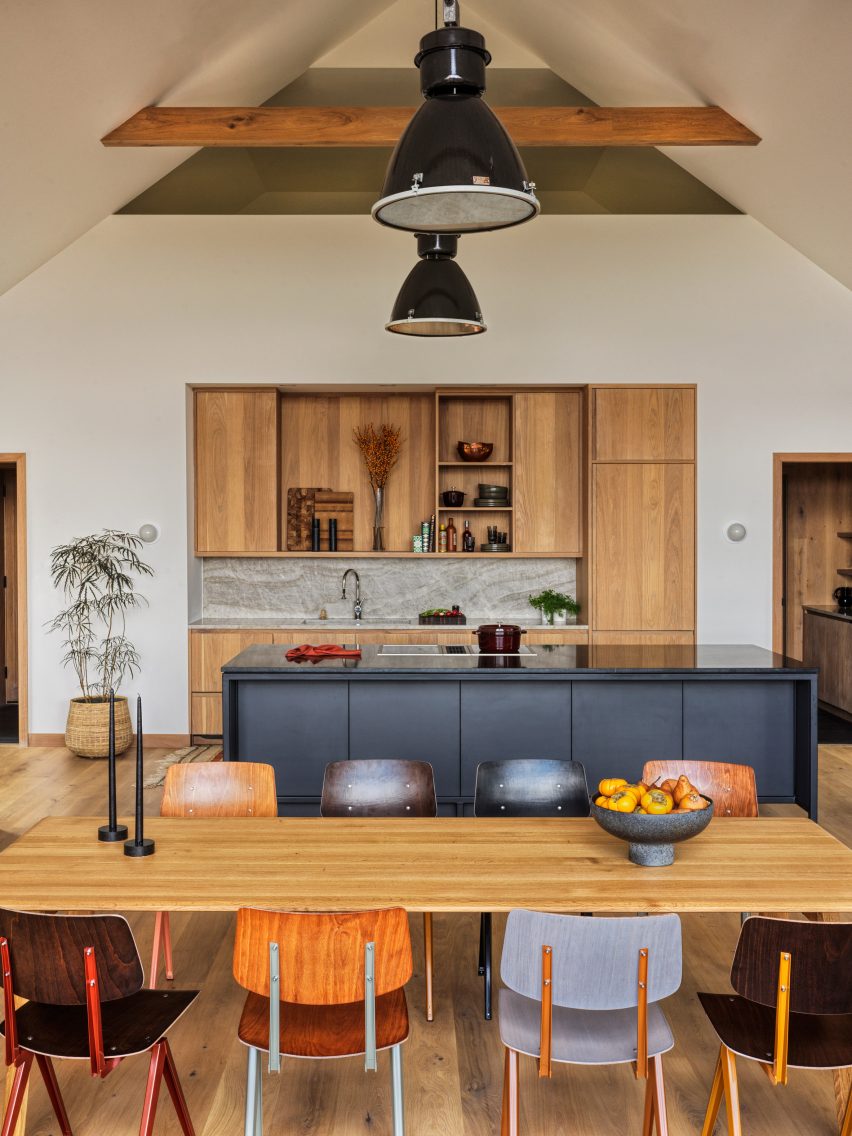 Otevřená kuchyně a jídelna s dřevěným stolem, barevnými jídelními židlemi, černým kuchyňským ostrůvkem, dřevěným kuchyňským úložným prostorem a odhalenou střešní konstrukcí