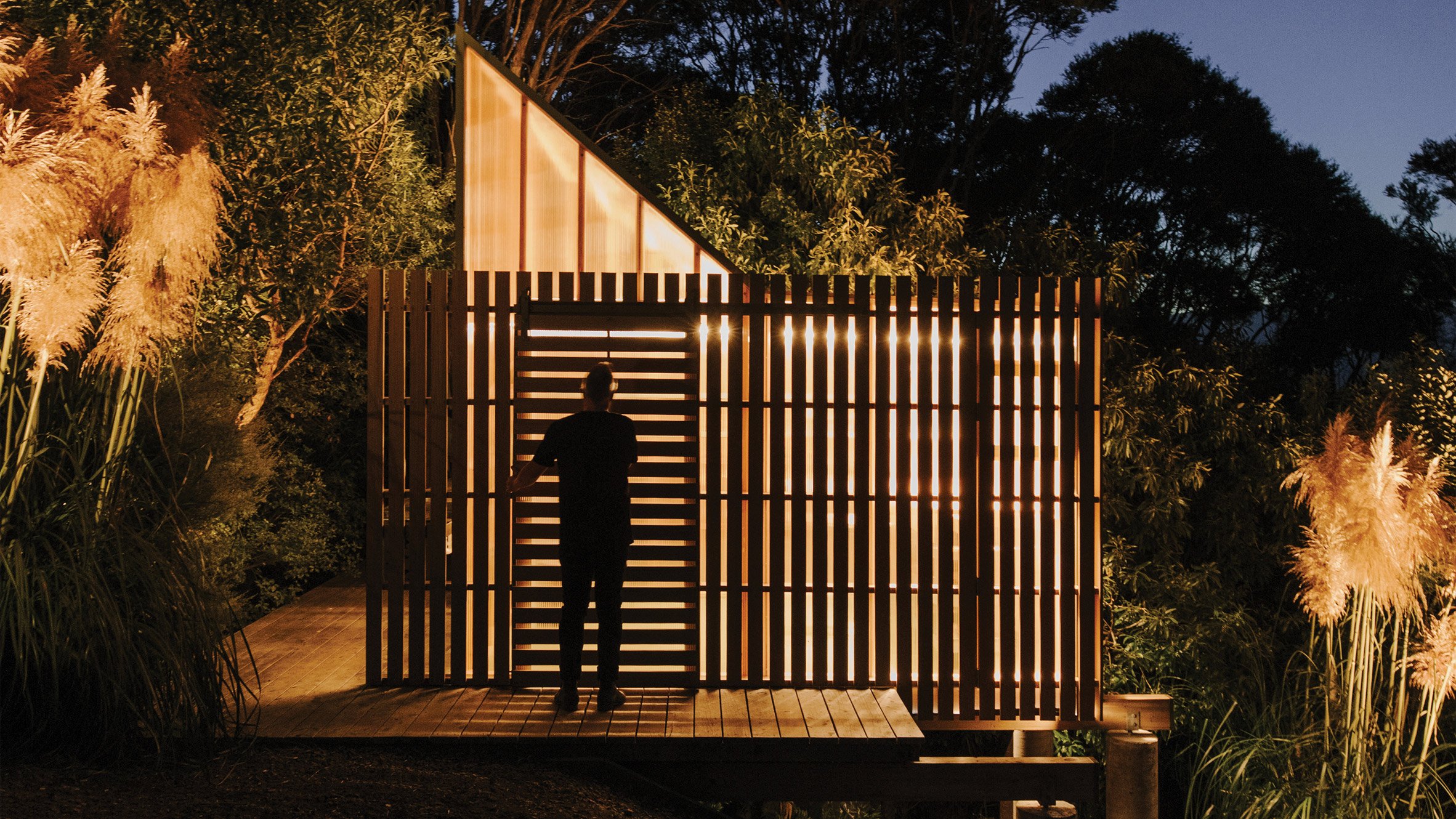 Silhouette of a person standing in front of a mono-pitched polycarbonate shed on a wooden deck shining light through timber slats on a slope in the New Zealand bush