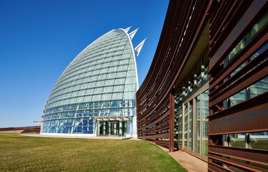 A curved, arched glass building next to a steel-clad building with a grassy lawn in front
