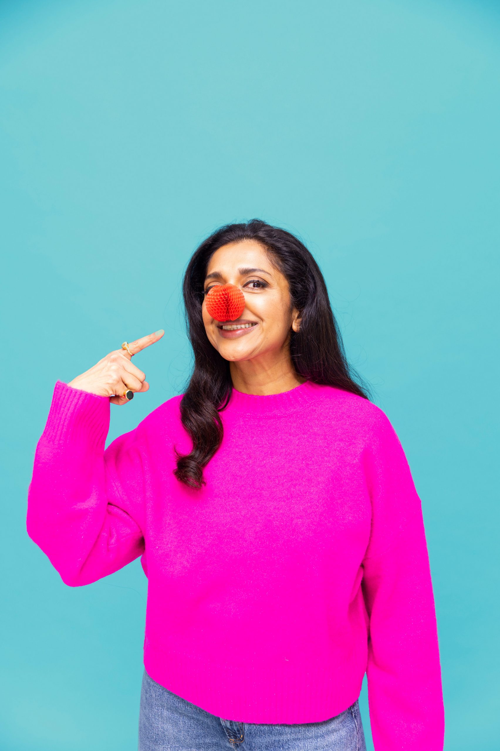 Photo of British actor Sindhu Vee proudly pointing to the Red Nose Day nose she is wearing