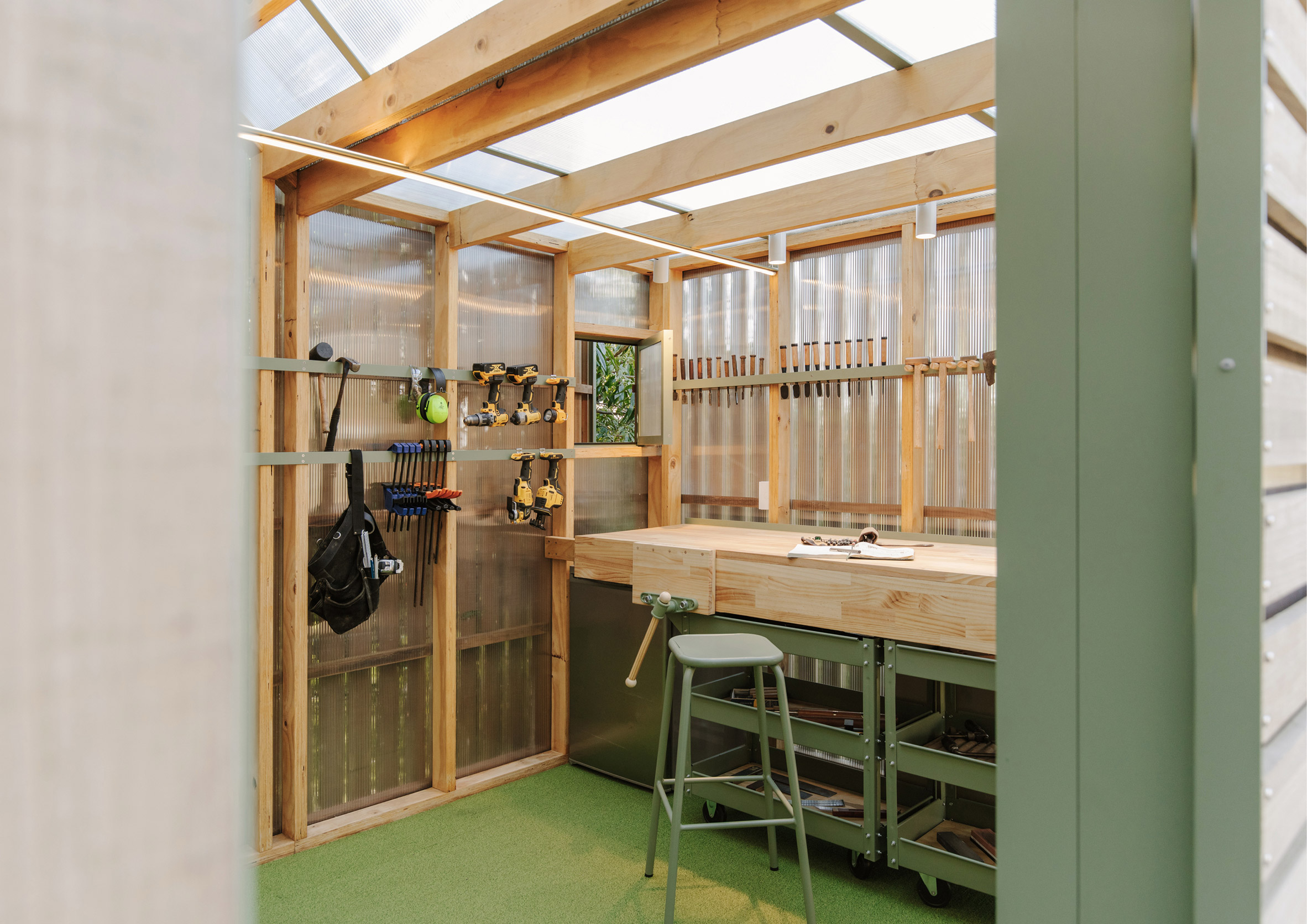 Interior of a timber and polycarbonate shed with a wood workbench, green floor, metal stool and storage