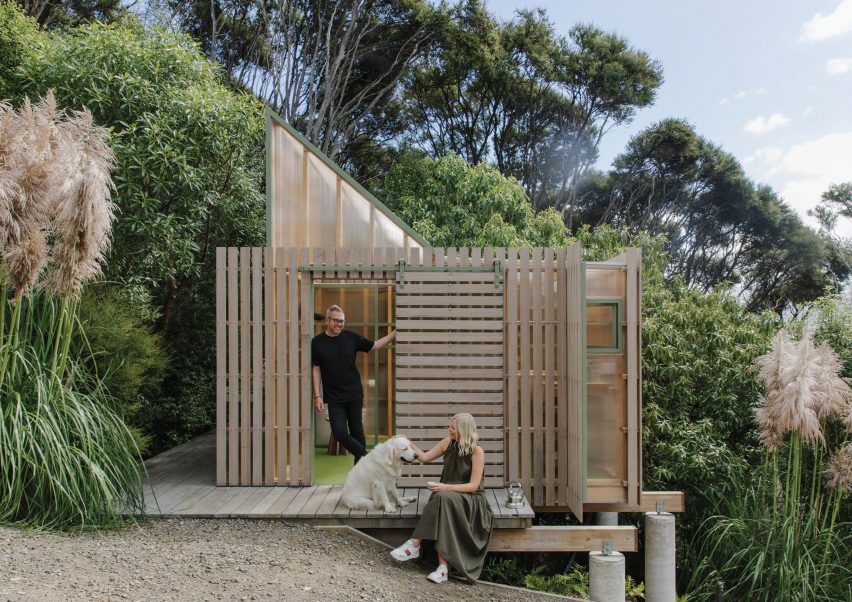 Two people and a dog on the decking of a mono-pitched shed covered in timber battens in a New Zealand bush