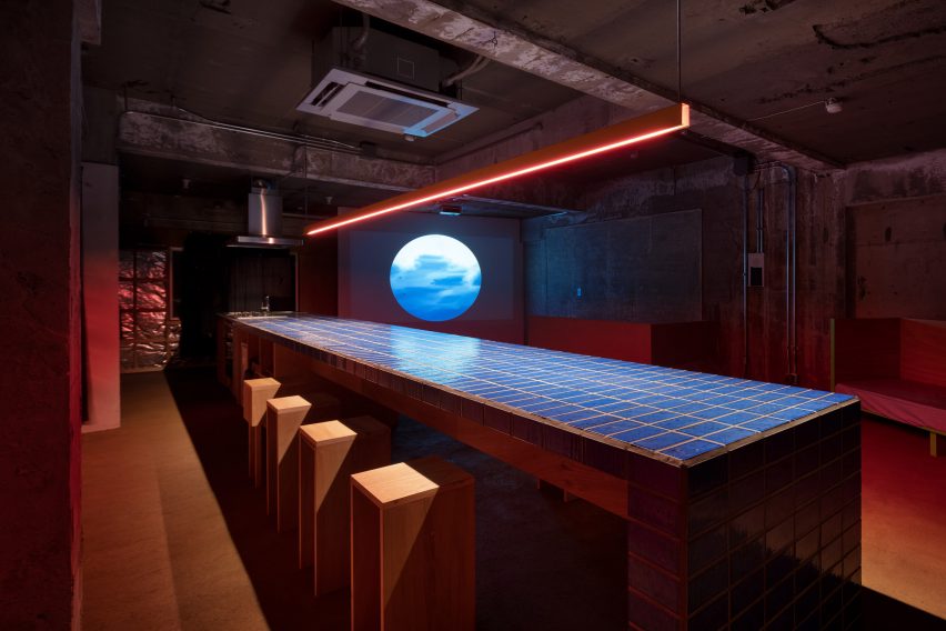 Long kitchen table topped with blue tiles with red strip lighting overhead in an unfinished industrial apartment