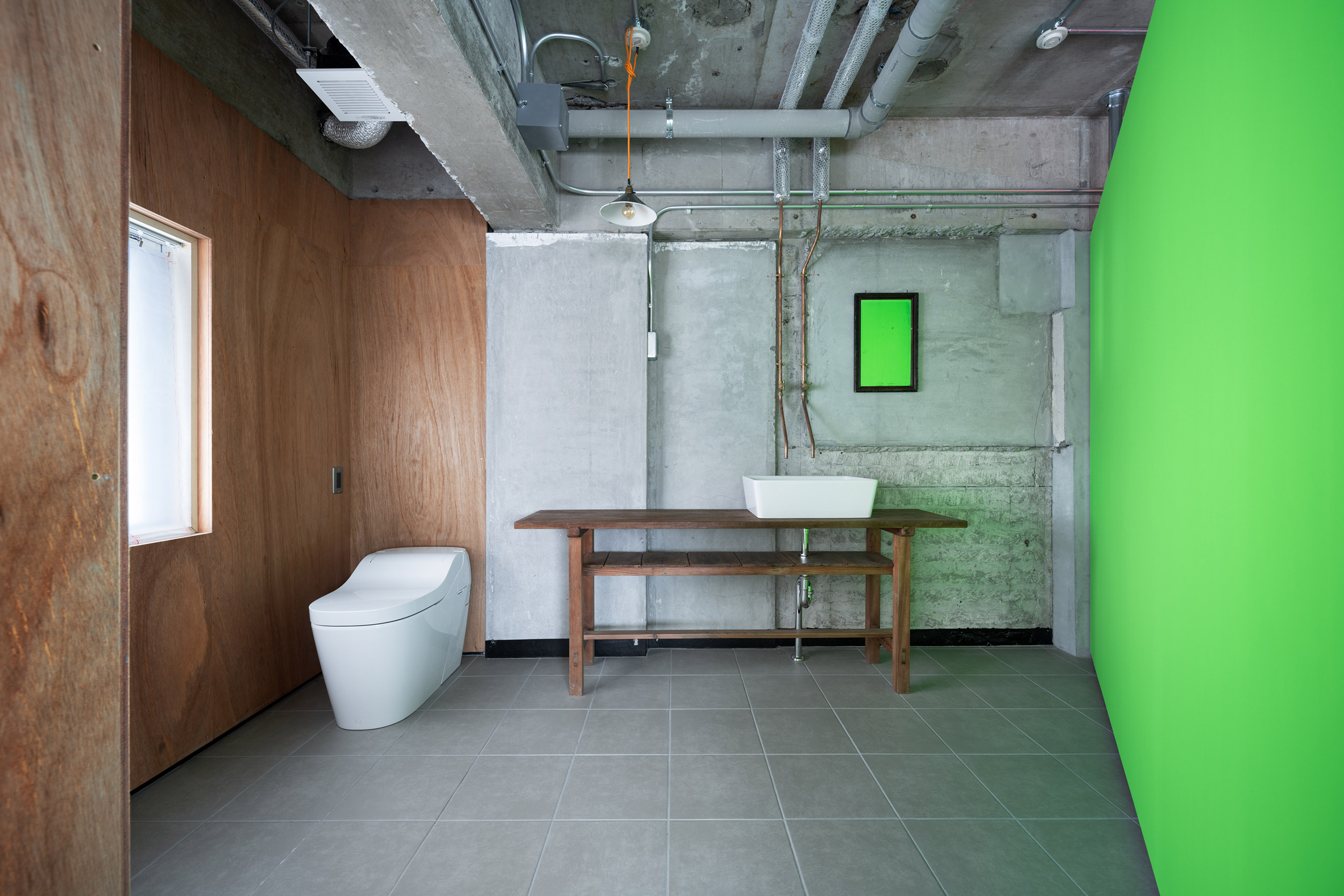 Toilet and sink on a wooden bench in an industrial bathroom with fluorescent green wall by Ab Rogers Design