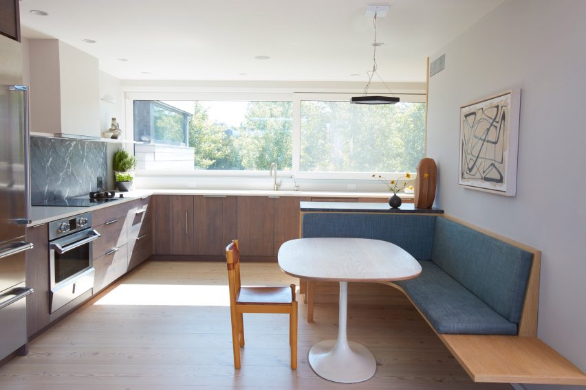 Interiér kuchyně se skříňkami z tmavého dřeva podél dvou kolmých stěn a dřevěným lavicovým sezením u bílého stolu