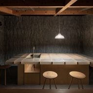A dark kitchen space with textured stone walls, timber frame roof and large central island with a sink lit by a pendant light