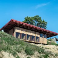 A guesthouse with large windows, wood walls and an overhanging red steel roof on a hillside
