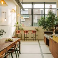 Communal kitchen and dining area with white walls and floors, large windows in dark metal frames, wood kitchen units, wood dining table and pink bar stools by The Mint List