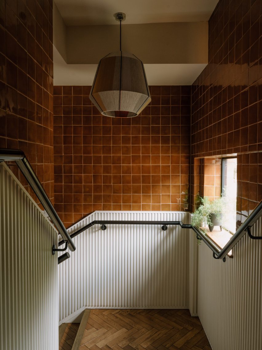 Escalera con paredes de azulejos marrones, paneles blancos y lámpara colgante de vidrio en el pasillo de The Mint List