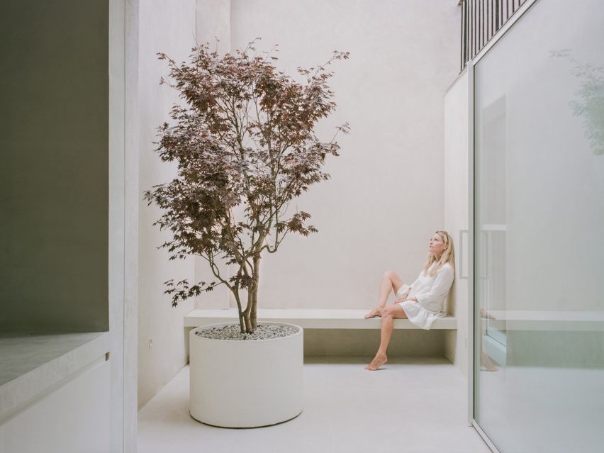 Un espacio interior con una puerta de vidrio pivotante que da a un patio exterior blanco con un banco y un árbol en una maceta
